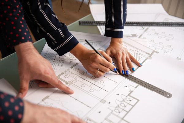 Two people reviewing a house floor plan with a pencil and ruler on a drafting table